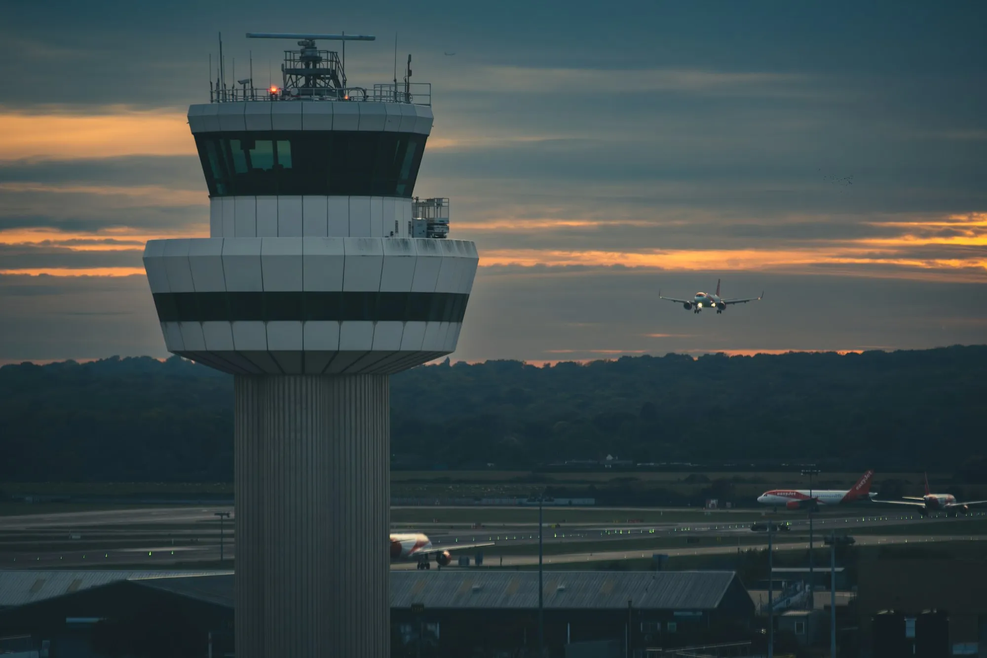 Torre de control de un aeropuerto al atardecer, con un avión en aproximación.